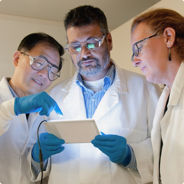 Three scientists in lab coats and gloves reviewing data on a tablet together.