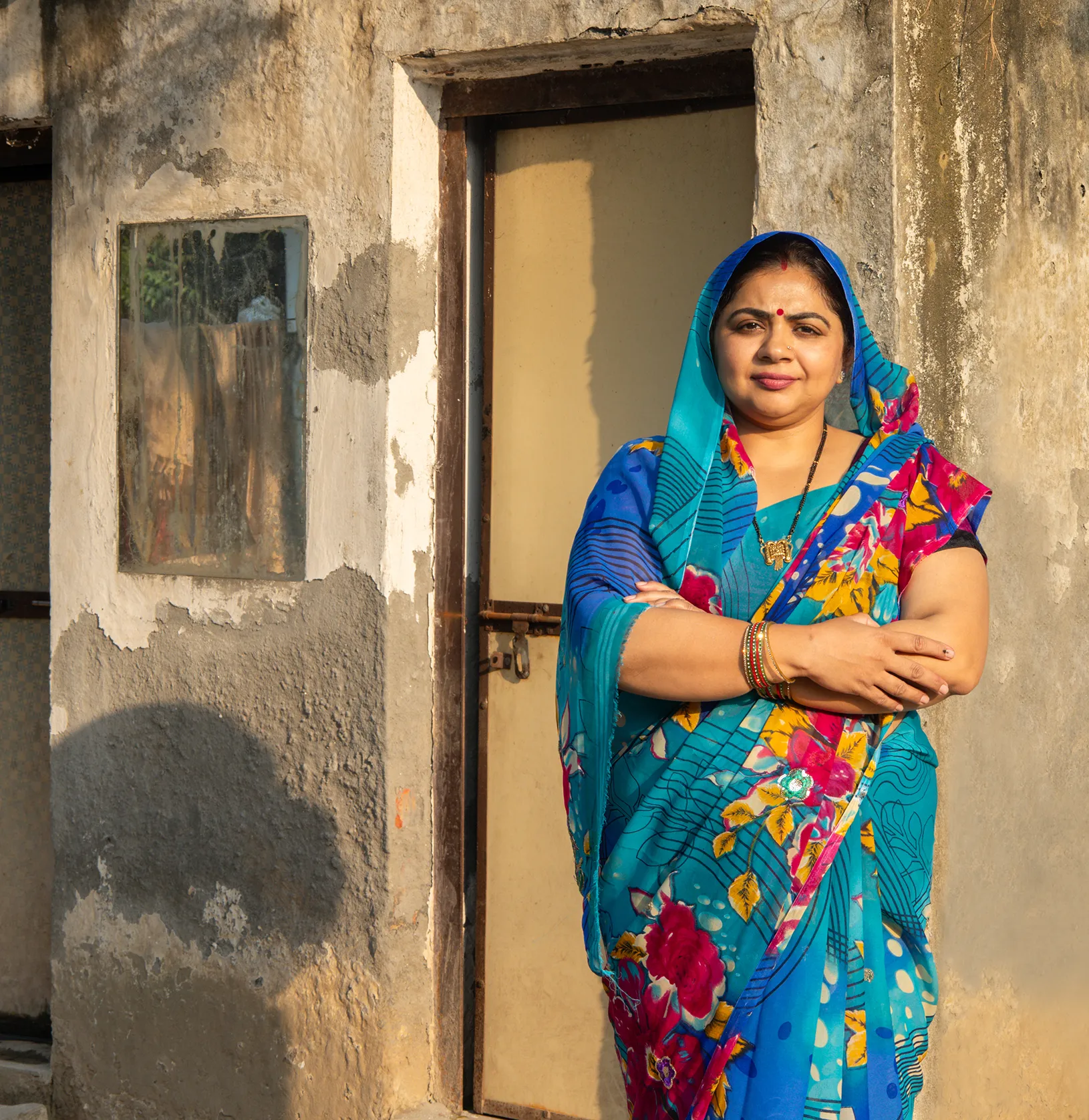 A woman in a blue and pink floral sari stands with arms crossed in a doorway of a weathered stone building, lit by warm sunlight.